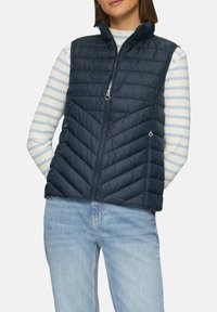 Visa en duvet bleu marine avec un matelassage en chevrons, un col haut et une fermeture éclair à l'avant. Associée à une chemise à rayures à manches longues en dessous.