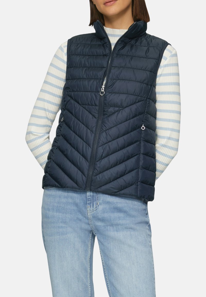 Visa en duvet bleu marine avec un matelassage en chevrons, un col haut et une fermeture éclair à l'avant. Associée à une chemise à rayures à manches longues en dessous.