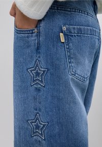 Jeans de mezclilla azul claro con dos diseños de estrellas bordadas en hilo de contraste en el lateral, con una etiqueta en el bolsillo trasero.