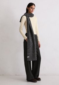 Longue écharpe grise au tricot texturé drapée sur un haut beige clair, associée à un pantalon noir à jambes larges et des chaussures à enfiler marron foncé.