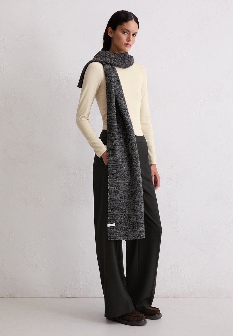 Longue écharpe grise au tricot texturé drapée sur un haut beige clair, associée à un pantalon noir à jambes larges et des chaussures à enfiler marron foncé.