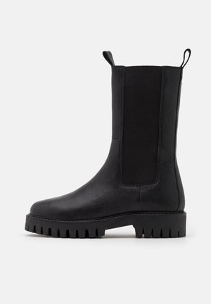 Botte noire en cuir mi-mollet avec semelle épaisse, panneau latéral élastique et languettes à l'avant et à l'arrière sur fond blanc uni.