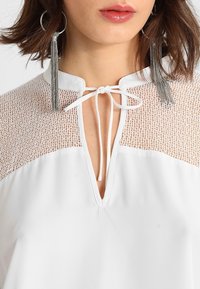 Vitt blus med en genomskinlig spetspanel på axlarna, med knytning vid halsringningen och en mjuk tygstruktur. Silverörhängen med tofsar.