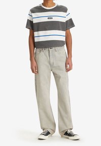 Grå och vit randig T-shirt med blå detaljer, svart logga, kombinerad med ljus tvättade baggy jeans och svarta sneakers. Avslappnad outfit visas.