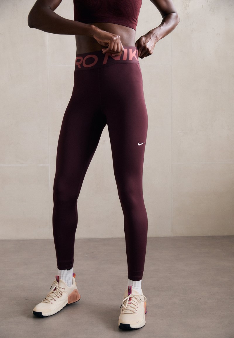 Leggings de sport bordeaux avec une taille brandée, texture lisse et un petit logo blanc sur la jambe gauche. Portés avec des baskets beiges et des chaussettes blanches.