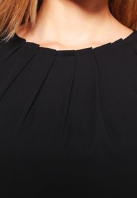 Blouse noire avec un design plissé autour de l'encolure. Texture lisse, tissu doux et détail de col rond sont visibles.