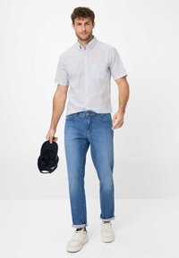 Camicia a maniche corte con motivo azzurro chiaro, abbinata a jeans blu medio e sneaker bianchi. Tiene un cappellino nero nella mano sinistra.