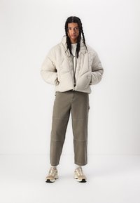 Veste matelassée beige clair avec col haut, portée par-dessus un sweat gris, associée à un pantalon cargo olive et des baskets beiges.