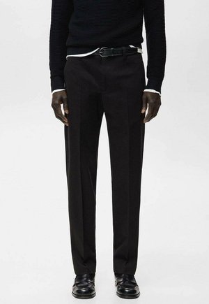 Mand iført sorte habitbukser, sort bælte, sorte loafers og sort tekstureret sweater over hvid skjorte står mod en ensfarvet baggrund.