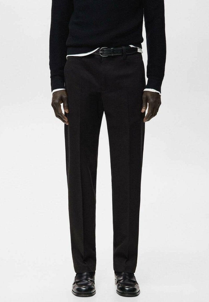 Homme portant un pantalon de costume noir, une ceinture noire, des mocassins noirs et un pull texturé noir par-dessus une chemise blanche, debout devant un fond uni.