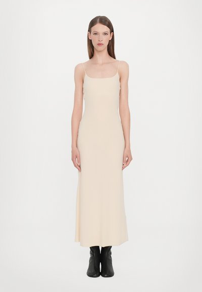 Beige lange slipdress met dunne spaghettibandjes, strak lijfje en vloeiende rok, gecombineerd met kniehoge zwarte laarzen. Soepele textuur.