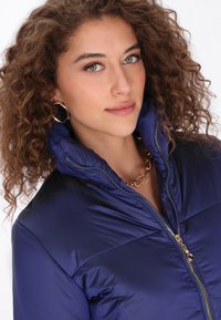 Jachetă pufică bleumarin cu guler înalt, finisaj lucios și fermoar auriu. Purată cu un colier din lanț auriu, având bucle texturate în păr.