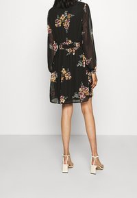 Černé květinové šaty s dlouhými průhlednými rukávy, zúženým pasem a volnou sukní. Obsahují jasné květinové vzory v žluté a burgundské barvě.