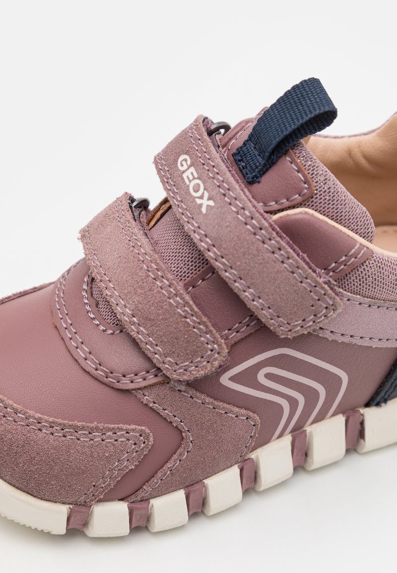 Sneakers in pelle rosa con due fascette in velcro, dettagli testurizzati e una suola in gomma bianca con una presa a motivo. Nome del marchio "GEOX" visibile.