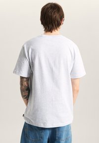 Grå bomullströja med korta ärmar och rund halsringning. Har en jämn textur och en avslappnad passform. Synlig tatuering på vänster arm.