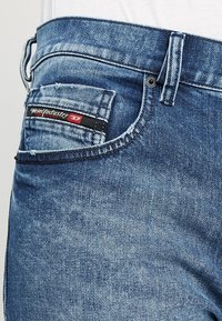 Jeans de denim azul claro con una textura ligeramente desgastada. Presenta un bolsillo trasero con una etiqueta negra y roja, un solo lazo para cinturón en la cinturilla y cremallera.