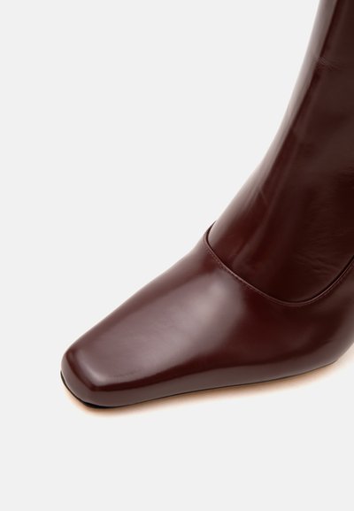 Botte en cuir marron foncé brillant avec un bout carré et une finition lisse sur un fond clair uni.
