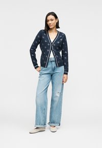 Μπλε ναυτικό ζακέτα με λευκή επένδυση και κεντήματα με λουλούδια, σε συνδυασμό με ανοιχτό μπλε τζιν παντελόνια φαρδιάς γραμμής. Το μοντέλο στέκεται με τα χέρια στη μέση, φορώντας μπεζ παπούτσια.