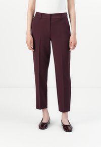 Žena v úzkých, ke kotníkům sahajících burgundy kalhotách s předním skladem, doplněná o shodné lakované baleríny.