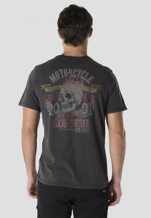 Hombre con una camiseta negra desgastada con una calavera en llamas y un gráfico con temática de motocicletas en la espalda, de pie frente a un fondo liso.