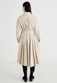 Béžový trench coat s opaskom, s prešívaným detailom na zadnej strane a dlhými rukávmi. V páre s čiernymi čižmami po kolená. Jednoduchý, klasický strih.