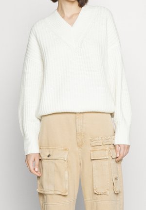 Pull blanc en maille avec col en V, bordures côtelées et motif texturé, associé à un pantalon cargo beige avec poches.