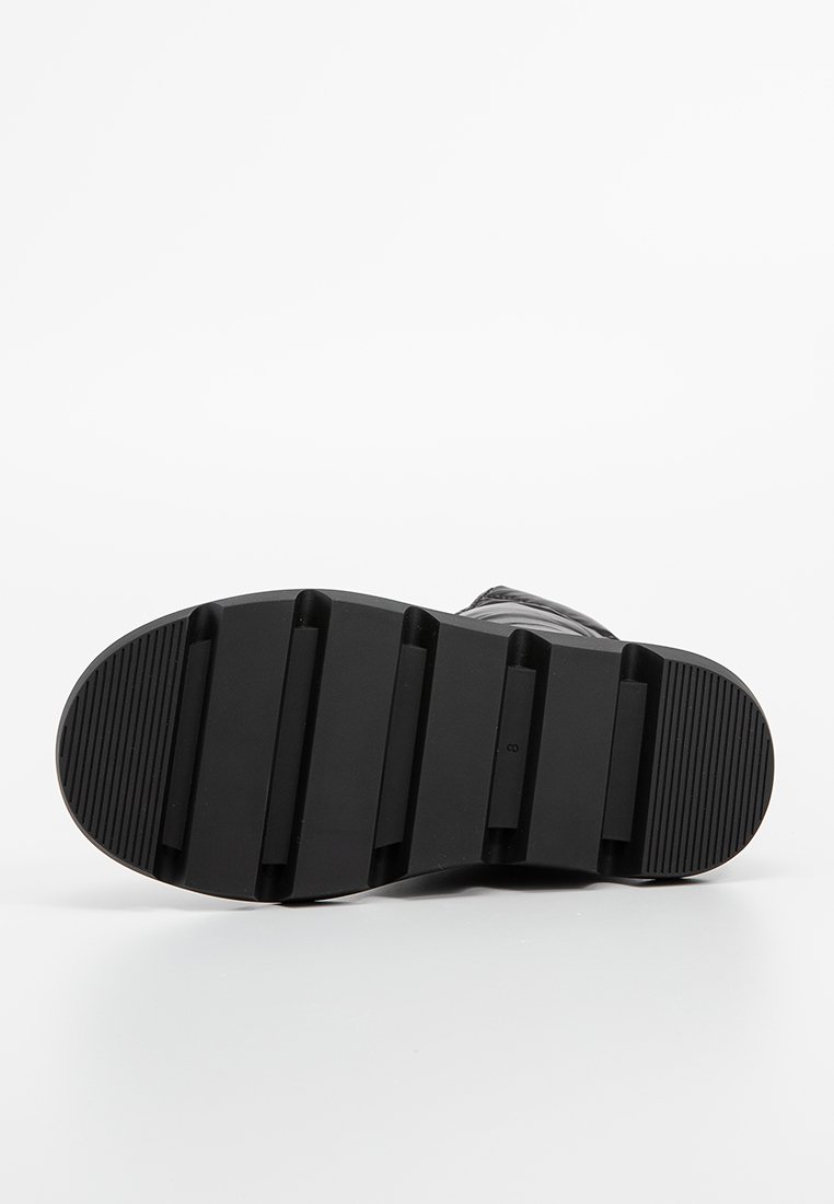 Suela de calzado negra con agarre texturizado, que presenta múltiples ranuras horizontales y verticales, puntera redondeada y material de goma resistente.