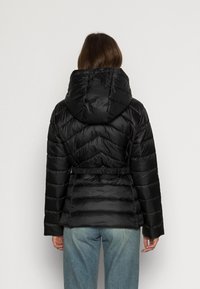 Giacca a vento nera con cappuccio, dotata di vita adattabile, texture trapuntata e bande elastiche sui polsini. Dettaglio di cuciture visibili su tutta la superficie.