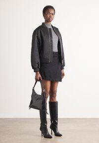 Giacca bomber nera sopra un dolcevita grigio aderente, abbinata a una mini gonna nera, stivali neri alti fino al ginocchio e una borsa nera strutturata.