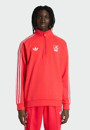 Mustanahuline inimene keskmise pikkusega dreadlocksidega, kandes punast Liverpool FC Adidas'i treeningülikonda, mille varrukatel ja pükstel on valged triibud.