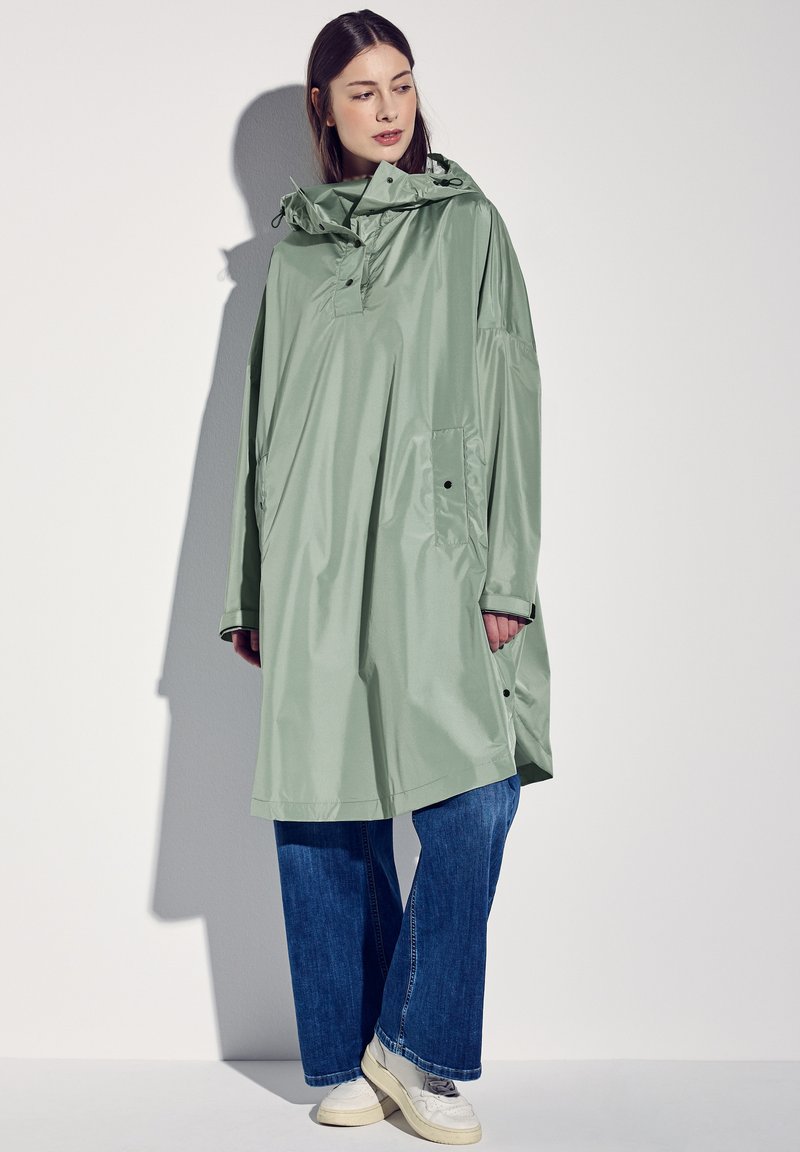 Jasnozielony, wodoodporny poncho z kapturem, bocznymi guzikami i dużą przednią kieszenią, noszony na luźnych niebieskich dżinsach i białych sneakersach.