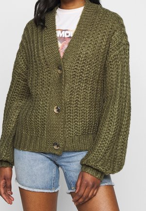 Femme portant un cardigan en maille épaisse vert olive avec des boutons, un t-shirt blanc à motif graphique et un short en denim bleu clair.
