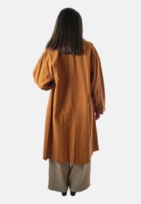 Een persoon met schouderlang donker haar draagt een lange bruine jas en brede beige broek, staande met de rug naar de camera op een plain witte achtergrond.