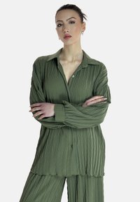 Chemise plissée vert olive avec un devant à boutons, un col et des manches longues, assortie avec un pantalon. Tissu texturé avec des lignes verticales.