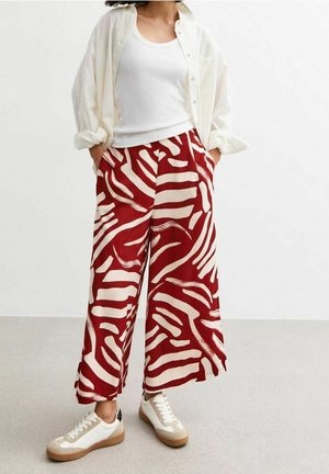 Personne portant un pantalon large à motifs abstraits rouges et blancs, un débardeur blanc, une chemise ouverte beige clair et des baskets blanches avec des semelles beiges.