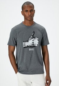 Camiseta de algodón gris con un gráfico en blanco y negro de una mano haciendo un gesto de aprobación y la palabra "Okey" debajo. Diseño de mangas cortas y cuello redondo.