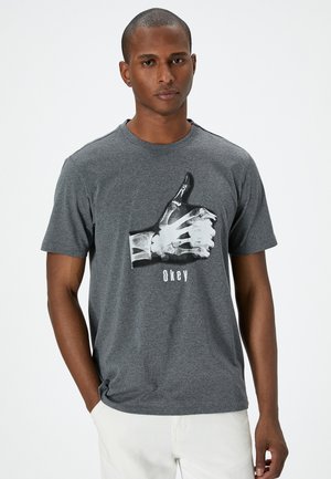 Camiseta de algodón gris con un gráfico en blanco y negro de una mano haciendo un gesto de aprobación y la palabra "Okey" debajo. Diseño de mangas cortas y cuello redondo.