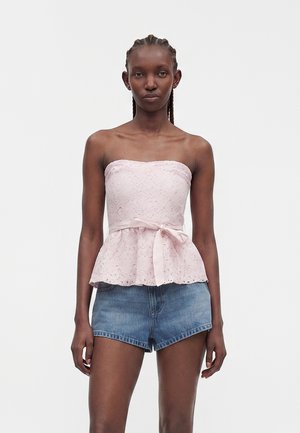 Kvinde med fletninger iført en lyserød blonder-top uden stropper med båndbælte og blå denimshorts, stående mod en ensfarvet baggrund.
