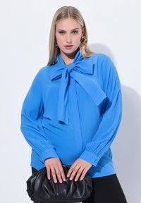 Blusa blu con un grande fiocco al collo, maniche lunghe e polsini abbottonati. Abbinata a una borsa a mano nera. Texture del tessuto liscia.