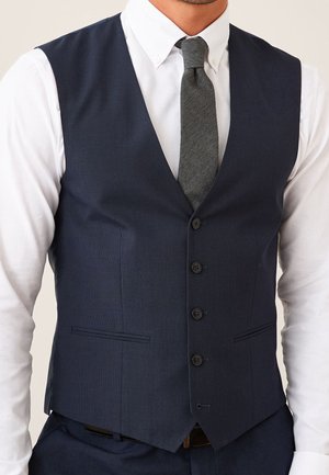Uomo che indossa un gilet blu navy abbottonato sopra una camicia bianca con una cravatta grigia con texture, mostrato dalle spalle alla vita.