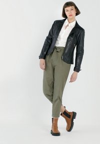 Zwarte leren motorjack, witte blouse, olijfgroene broek met strikceintuur, en bruine enkellaarzen met zwarte zolen. Glad textuur en aansluitend ontwerp.