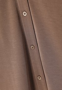 Camicia in tessuto marrone con texture, caratterizzata da bottoni chiari, che mostra un primo piano del colletto e dei dettagli dei bottoni.