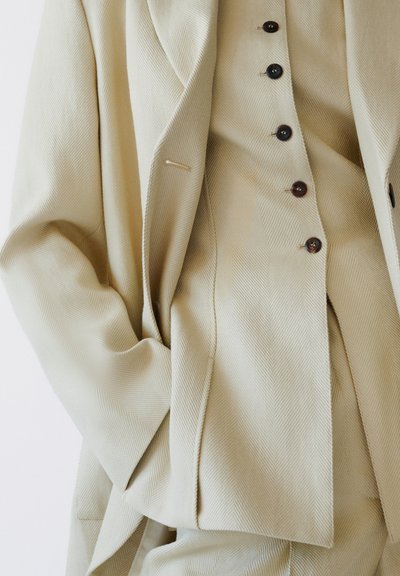 Μπεζ σακάκι με υφή και αντιστοιχισμένο γιλέκο με σκούρα κουμπιά, με το χέρι στην τσέπη του σακακιού, εμφανίζεται από το μέσο του κορμού μέχρι τους μηρούς.