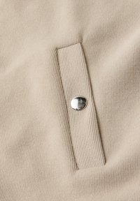 Gros plan sur un tissu beige côtelé avec une languette rectangulaire verticale fixée par un bouton pression argenté brillant.