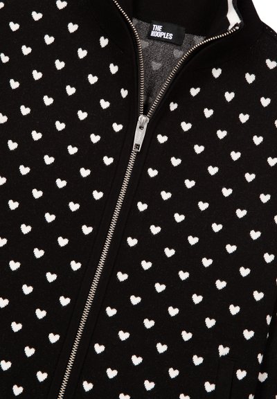 Cardigan noir zippé avec un motif de petits cœurs blancs partout. Confectionné en tissu doux, avec une fermeture éclair argentée et un col côtelé.