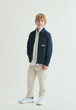 Blonder Junge steht vor hellem, einfarbigem Hintergrund. Er trägt eine marineblaue Jacke über einem weißen Reißverschluss-Pullover, beige Cargohosen und weiße Turnschuhe.