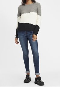 Pletený svetr s horizontálními barevnými bloky v šedé, bílé a černé. Spárován se štíhlými modrými džíny a černými botami na silné podrážce.