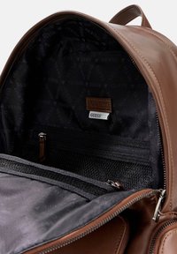 Intérieur d'un sac à dos en cuir brun doté d'un revêtement en tissu noir avec le motif "GUESS" répété, une poche zippée et une étiquette en cuir.