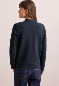 Felpa blu scuro, testurizzata, con motivo a spina di pesce, polsini e orlo a costine. Abbinate a jeans in denim scuro, viste da dietro.