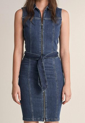 Ärmelloses dunkelblaues Jeanskleid mit einem Reißverschluss vorne und einem passenden Stoffgürtel, der in der Taille gebunden ist, getragen von einer stehenden Person.